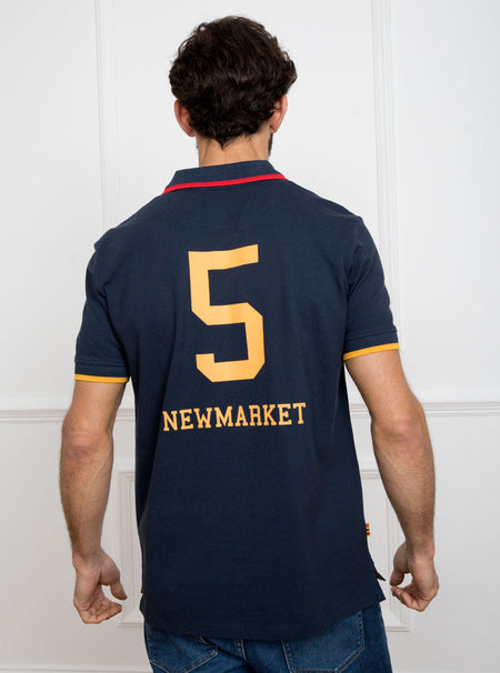 Man wearing navy polo shirt in 100% cotton with Newmarket 875 crest embroidered on the front and giant gold number 5 on the back with gold sleeve trimming and red collar trimming.